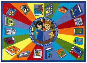 A childrens rug with an image of two kids reading the same book. The rug has different colors and books around the edge.
